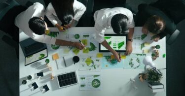 Vista superior de quatro pessoas em uma sala de reuniões. Na mesa, encontram-se papéis com gráficos e notebooks. Vista aérea de empresários trabalhando juntos para planejar e investir em sustentabilidade empresarial.