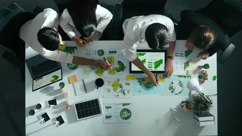 Vista superior de quatro pessoas em uma sala de reuniões. Na mesa, encontram-se papéis com gráficos e notebooks. Vista aérea de empresários trabalhando juntos para planejar e investir em sustentabilidade empresarial.